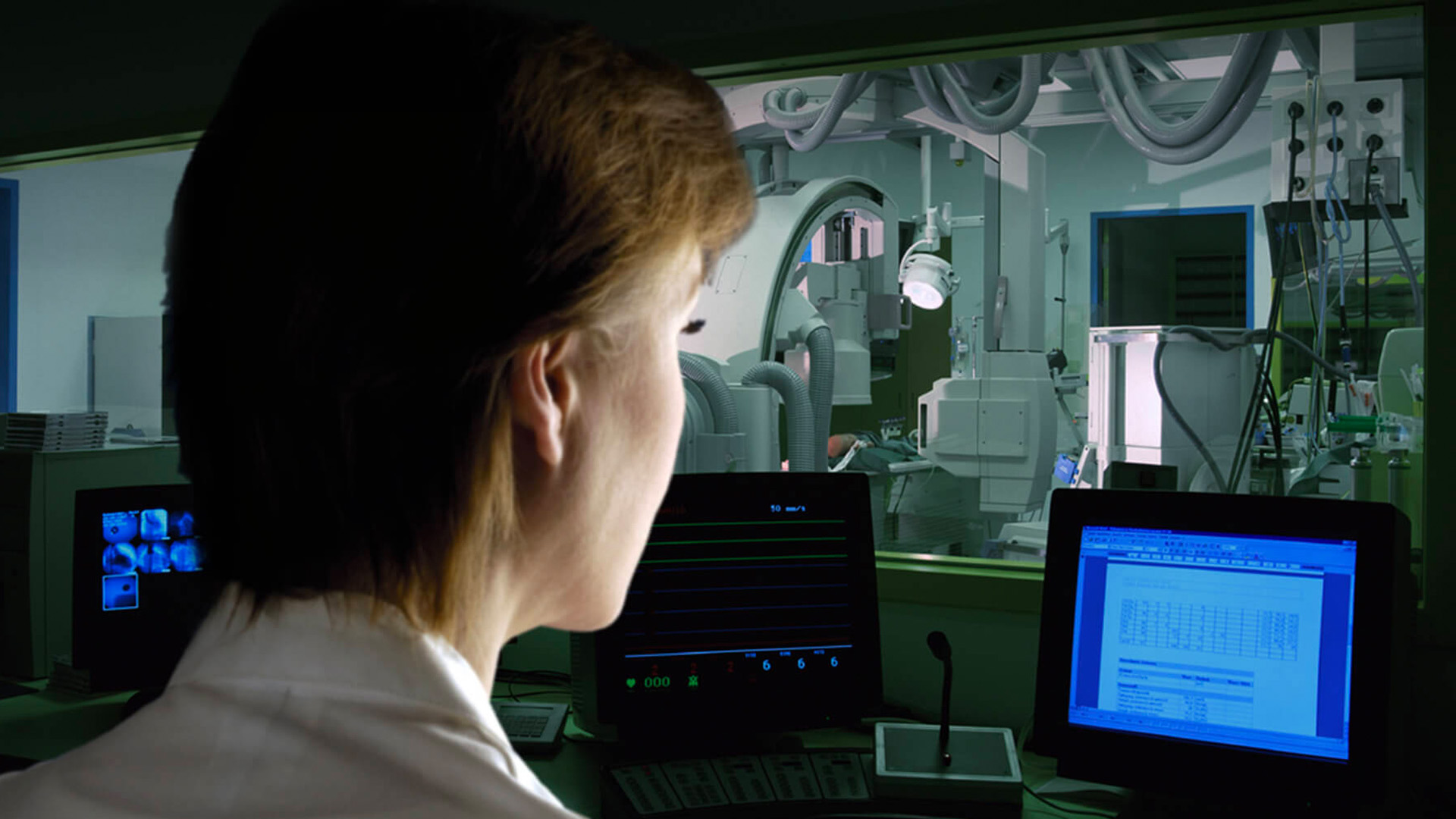 Ärztin beobachtet vom durch Strahlenschutzglas gesicherten Kontrollraum ein Röntgengerät