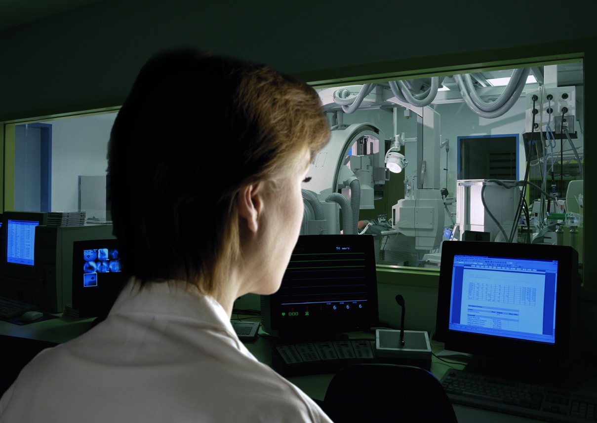 Ärztin beobachtet vom durch Strahlenschutzglas gesicherten Kontrollraum ein Röntgengerät