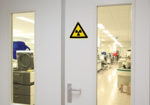 Zweiflügelige Strahlenschutztür mit Fenstern aus gelblichem Bleiglas, die einen Blick auf Röntgen-Geräte und andere radiologische Ausrüstung freigeben.